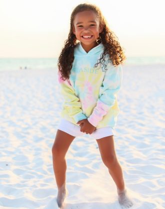 Smiling child wearing pastel tie-dye hoodie stands barefoot on white sandy beach at sunset.
