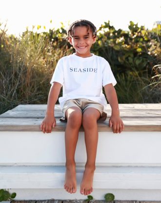 Smiling child wearing a “Seaside” shirt sitting barefoot on a wooden bench outdoors.