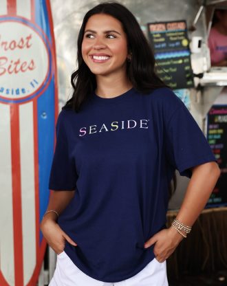 Smiling woman wearing navy “Seaside” shirt stands near colorful surfboard and food stand.