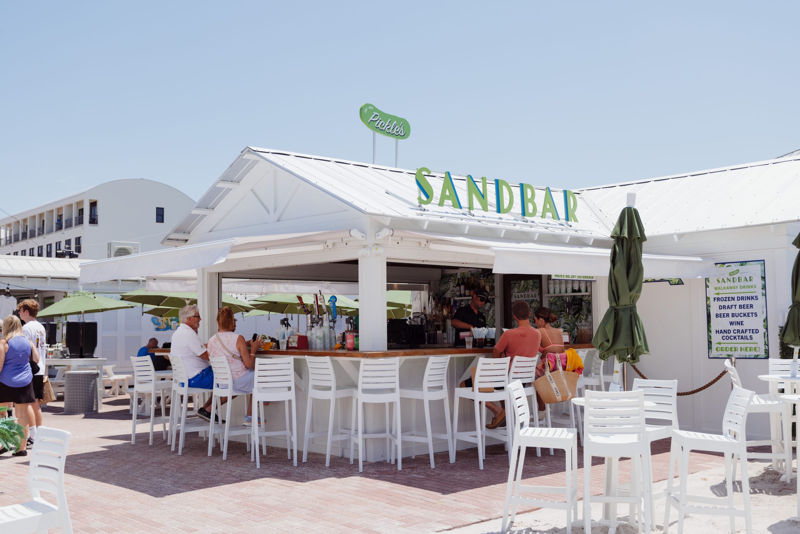 People relax at Pickle’s Sandbar, an open-air beach bar with white seating and green umbrellas.