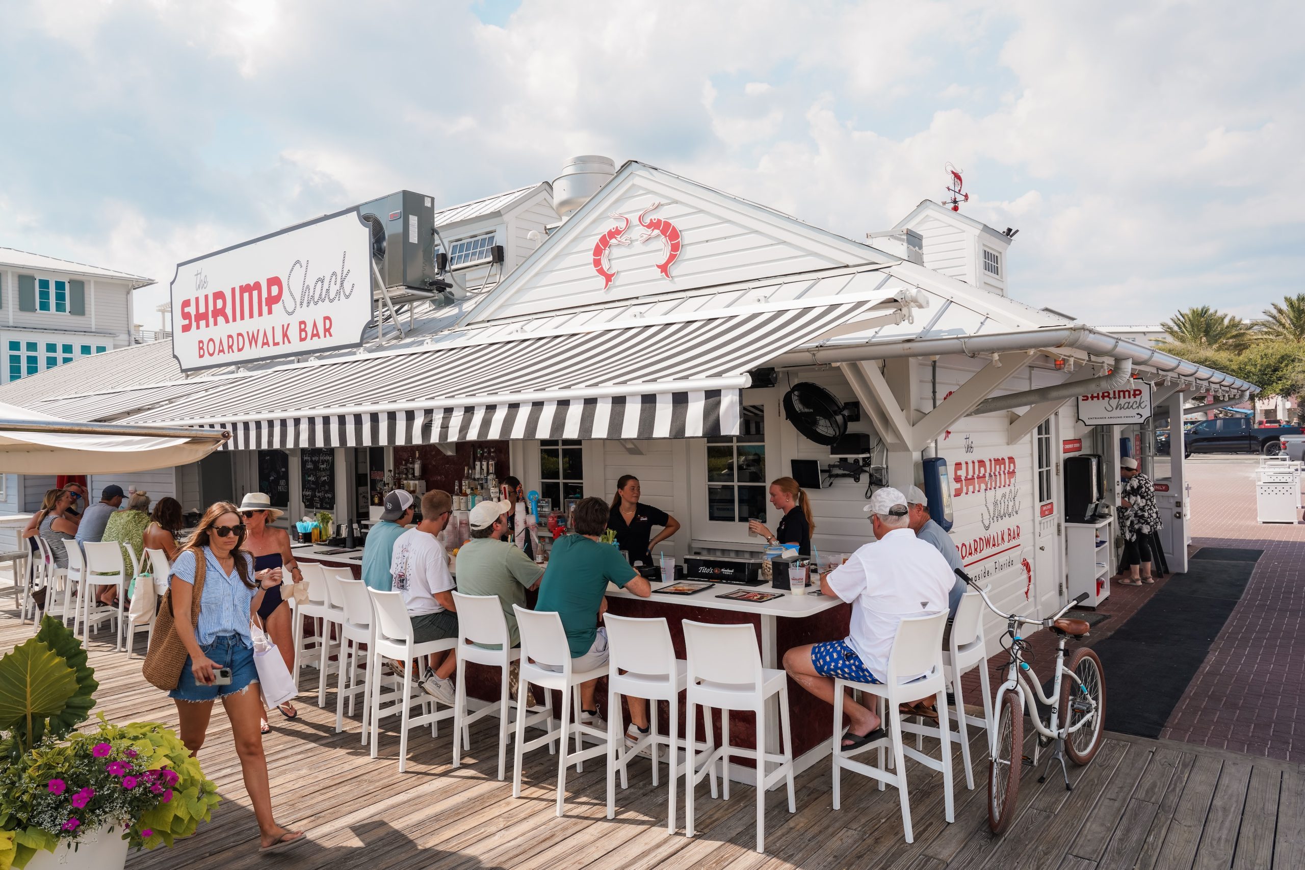 People sit at an outdoor bar under striped awnings at The Shrimp Shack Boardwalk Bar near beach.