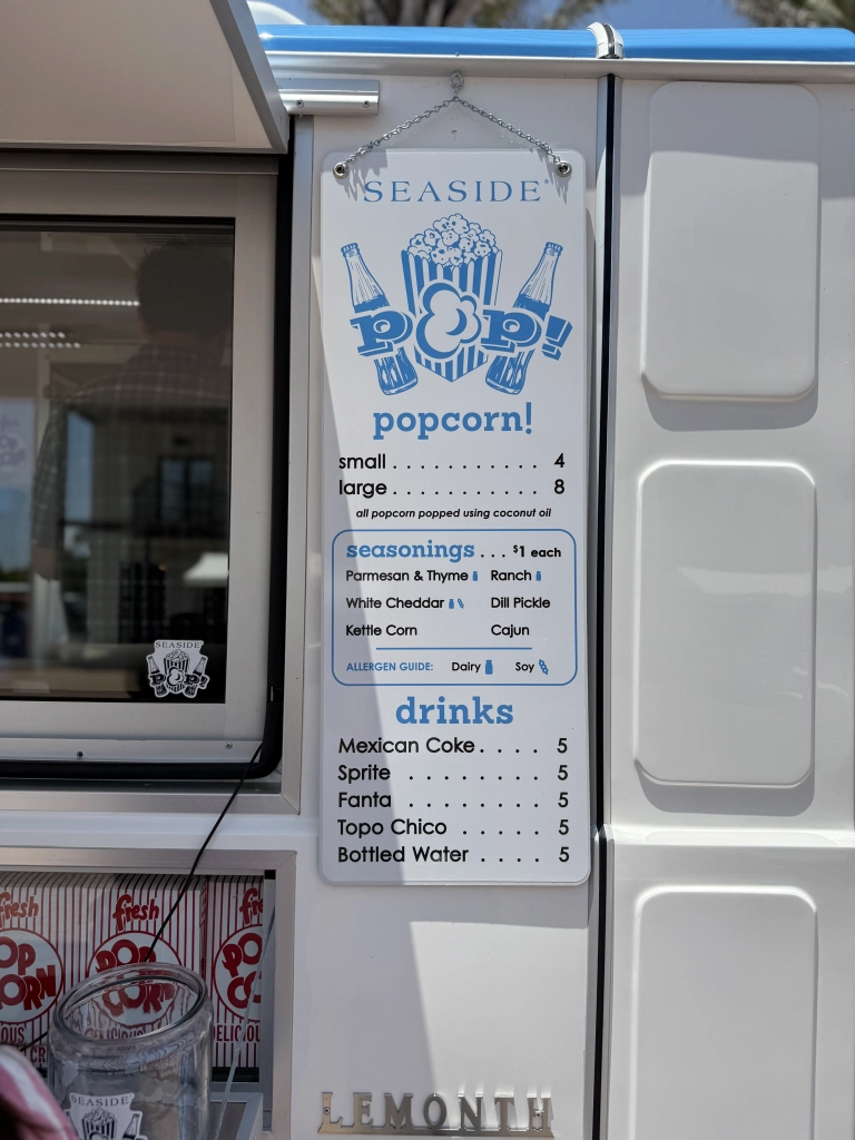 Menu sign on a seaside popcorn stand listing popcorn sizes, seasonings, and bottled drinks in blue and white.