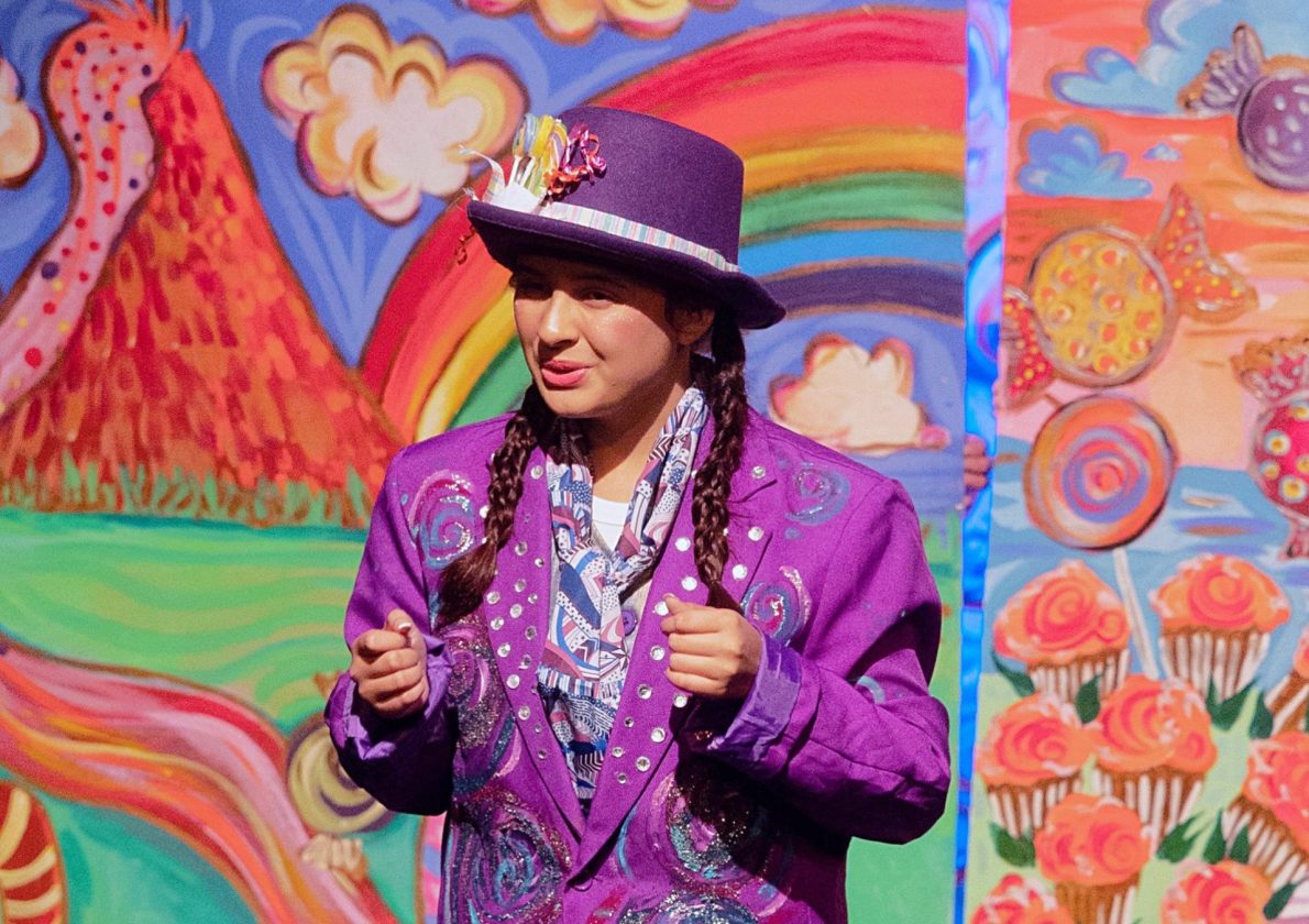 Performer in sparkling purple costume onstage before whimsical rainbow and candy backdrop.