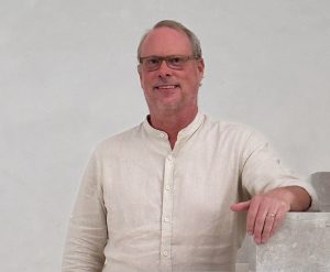 Smiling man wearing light linen shirt and glasses posing against neutral wall on staircase.