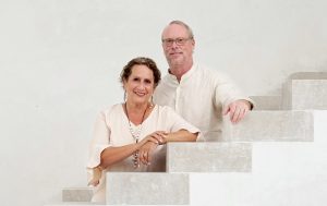 Man and woman wearing white clothing pose, leaning on modern white stairs with minimalist background.
