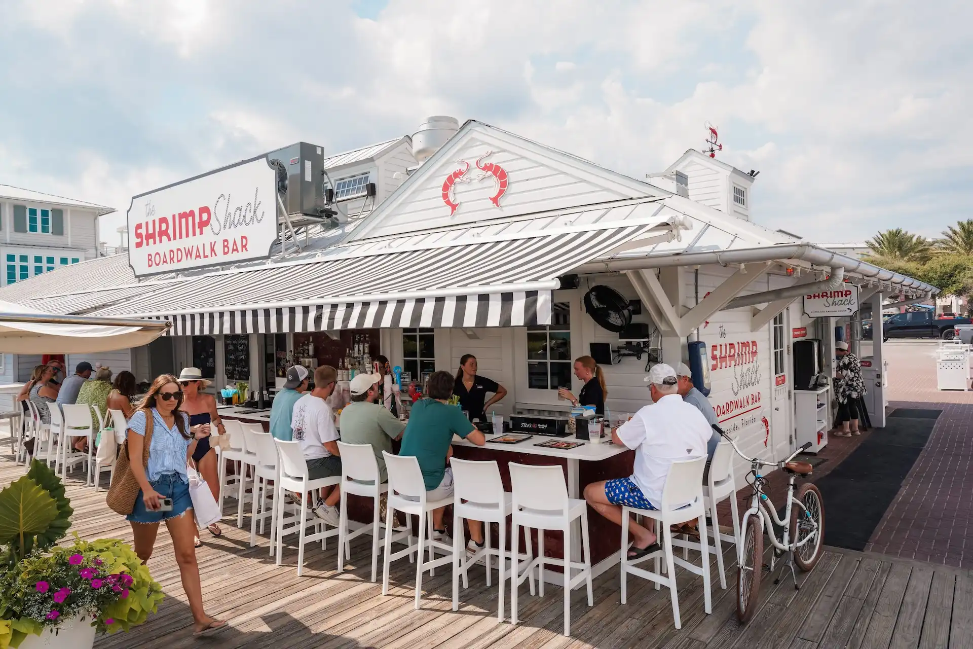 People sit at an outdoor bar under striped awnings at The Shrimp Shack Boardwalk Bar near beach.