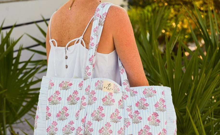 Woman in white sundress carries large floral tote bag over her shoulder, standing beside green palm plants.