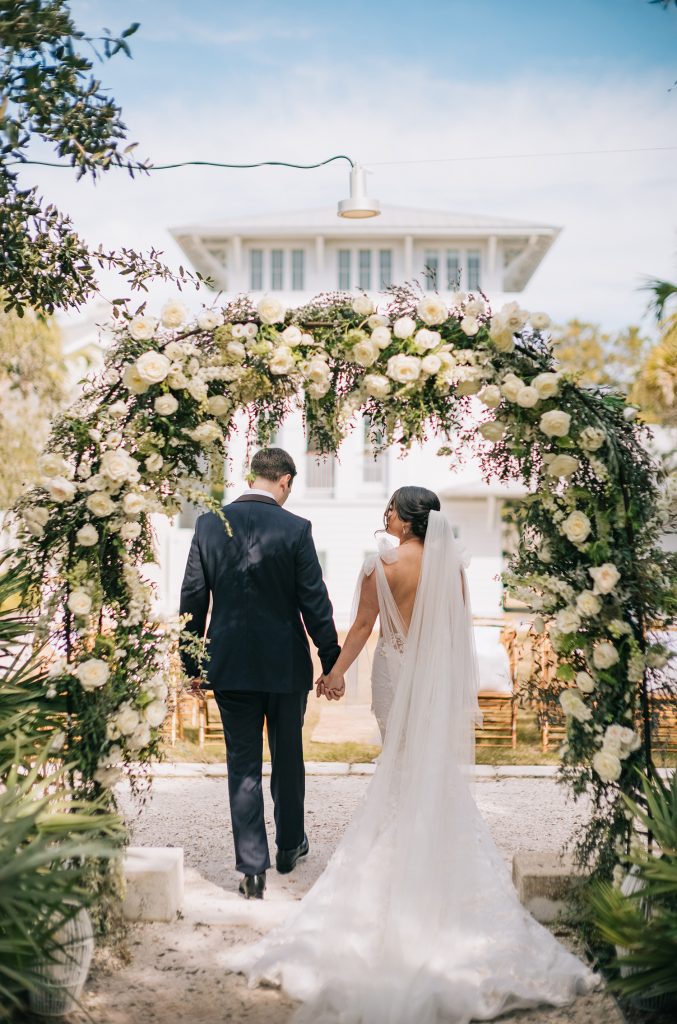 A bride and groom walk hand in hand beneath a lush floral arch at an outdoor wedding ceremony.