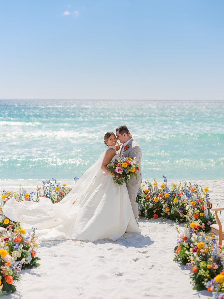 Bride and groom embracing on a sandy beach, framed by colorful flowers with turquoise ocean behind them.