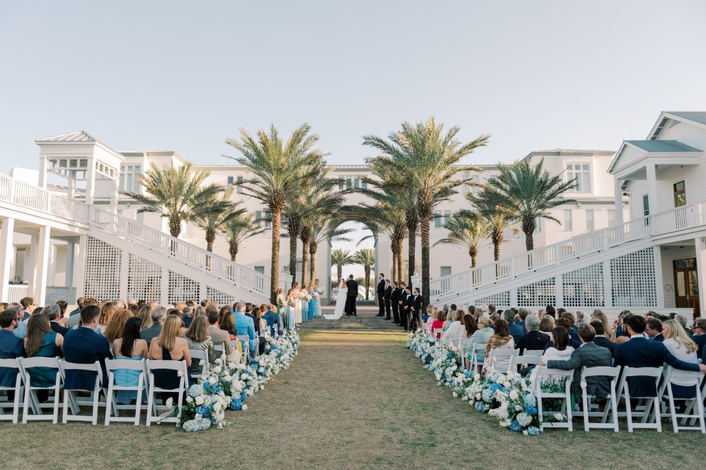 Outdoor wedding ceremony between palm-lined white buildings, guests seated along aisle of blue and white flowers.