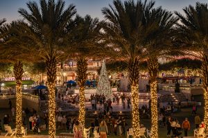 Palm trees wrapped in lights surrounding a festive outdoor plaza with people gathering around a lit holiday tree at dusk