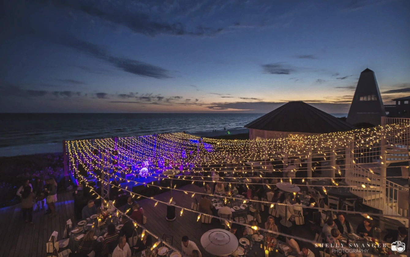 String lights illuminate outdoor beachside event at sunset, with guests dining under vibrant evening sky.