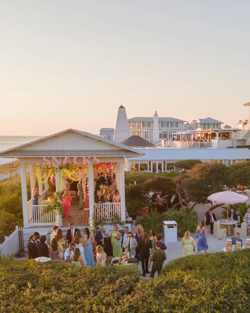 Guests mingling at an outdoor seaside reception near a pavilion, surrounded by coastal buildings at sunset.