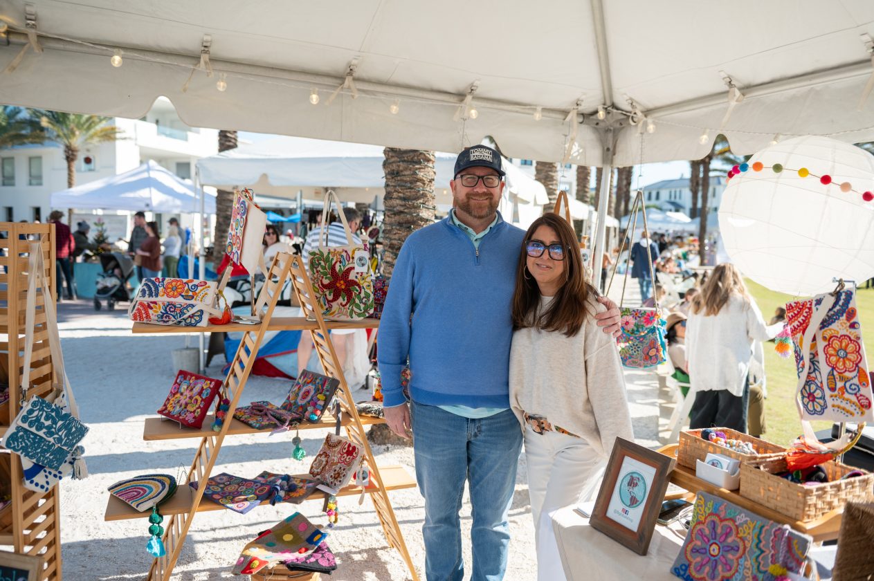 Couple stands inside market booth displaying colorful handmade bags and crafts at outdoor artisan fair.