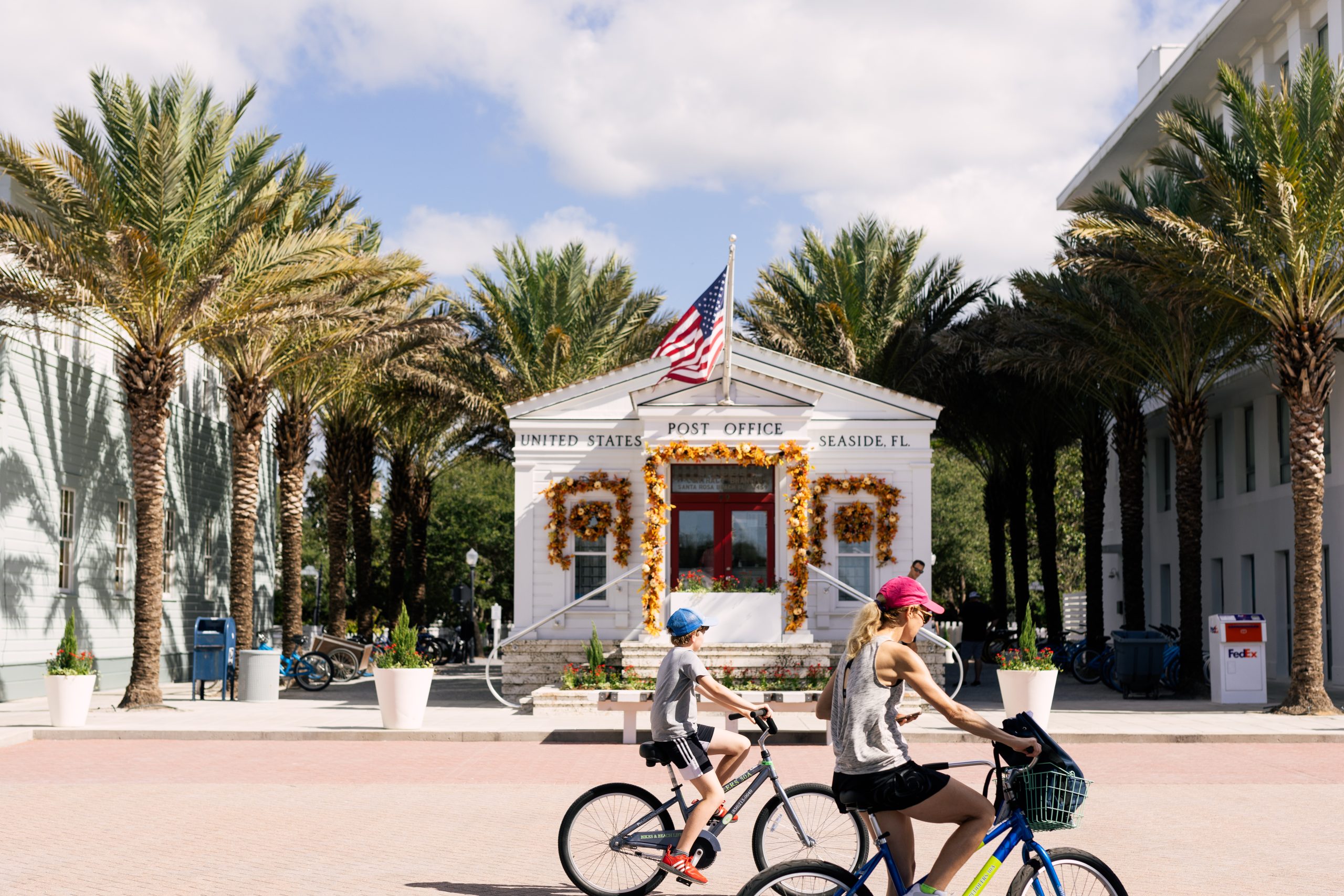 Two people ride bicycles past United States Post Office building decorated with garlands, palm trees, and flag.