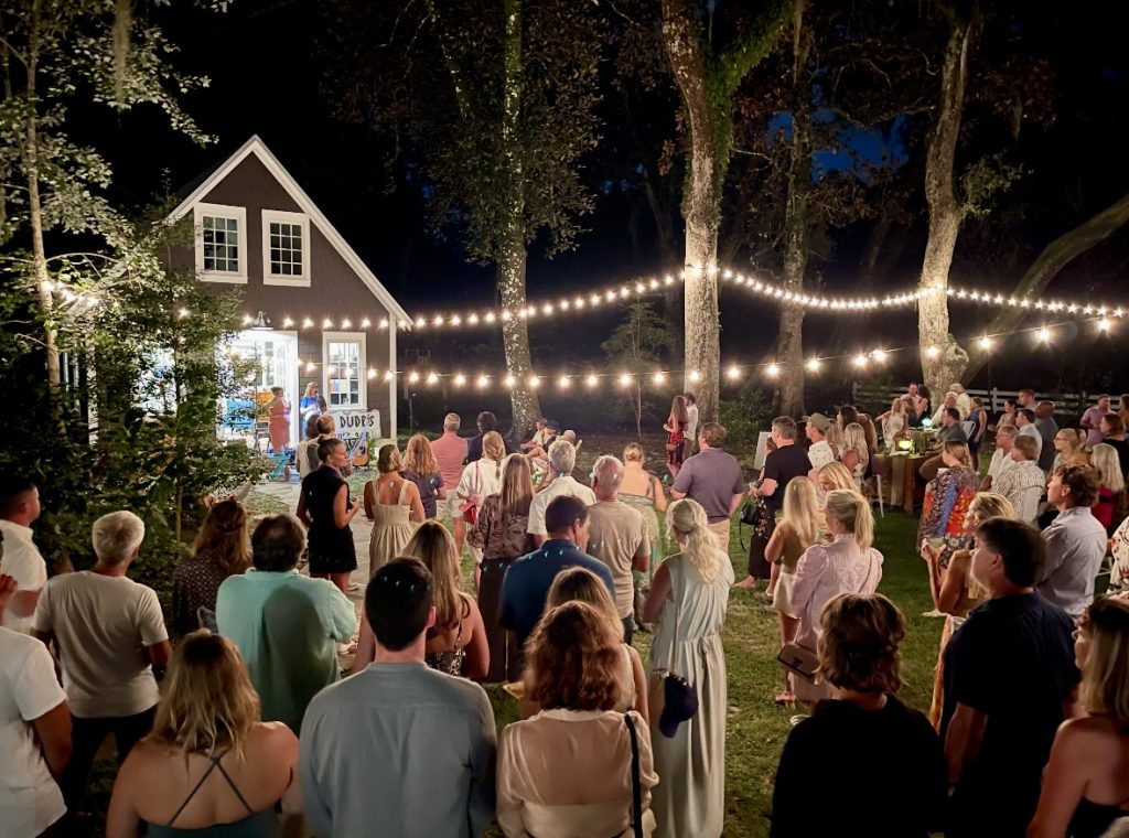 A crowd gathers outdoors at night beneath string lights, listening to speakers near a small building surrounded by trees.