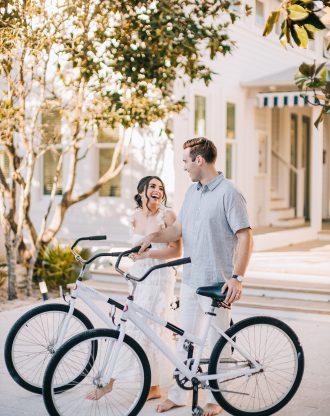 Smiling couple walking white bicycles outside a bright, sunlit coastal home.