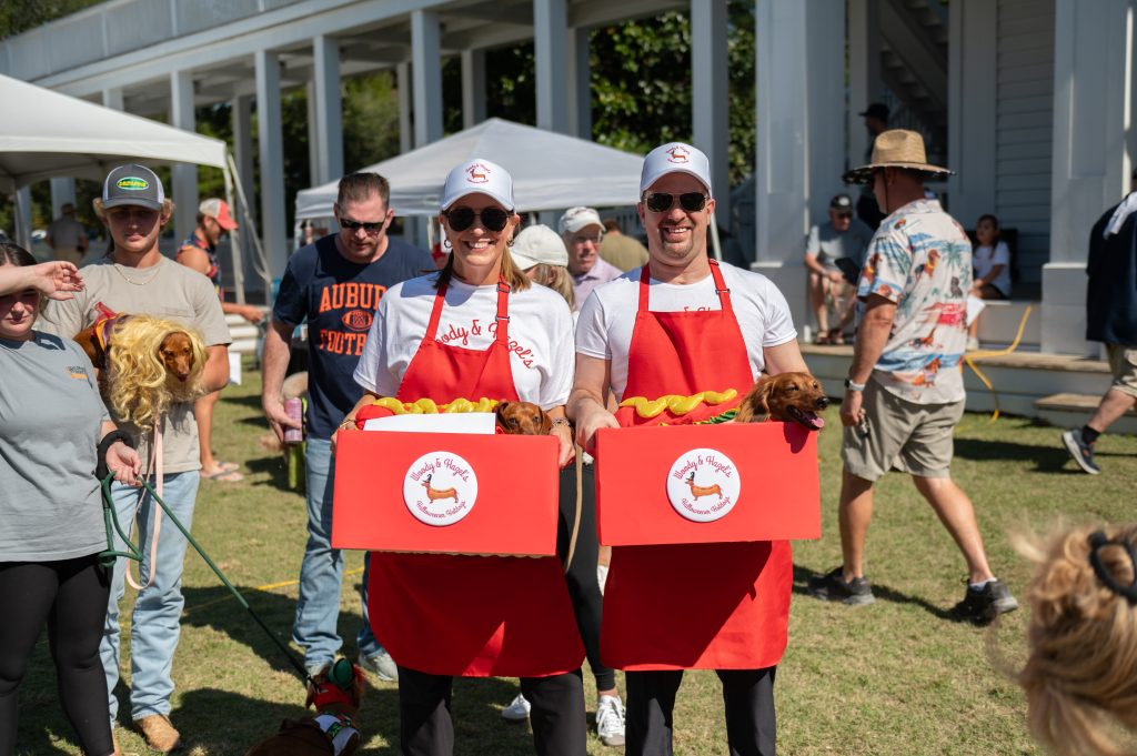Two people in red aprons holding boxes with dachshunds dressed as hot dogs at an outdoor event.
