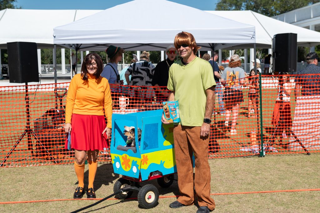 Two people dressed as Scooby-Doo characters pose beside a dog in a themed wagon outdoors.