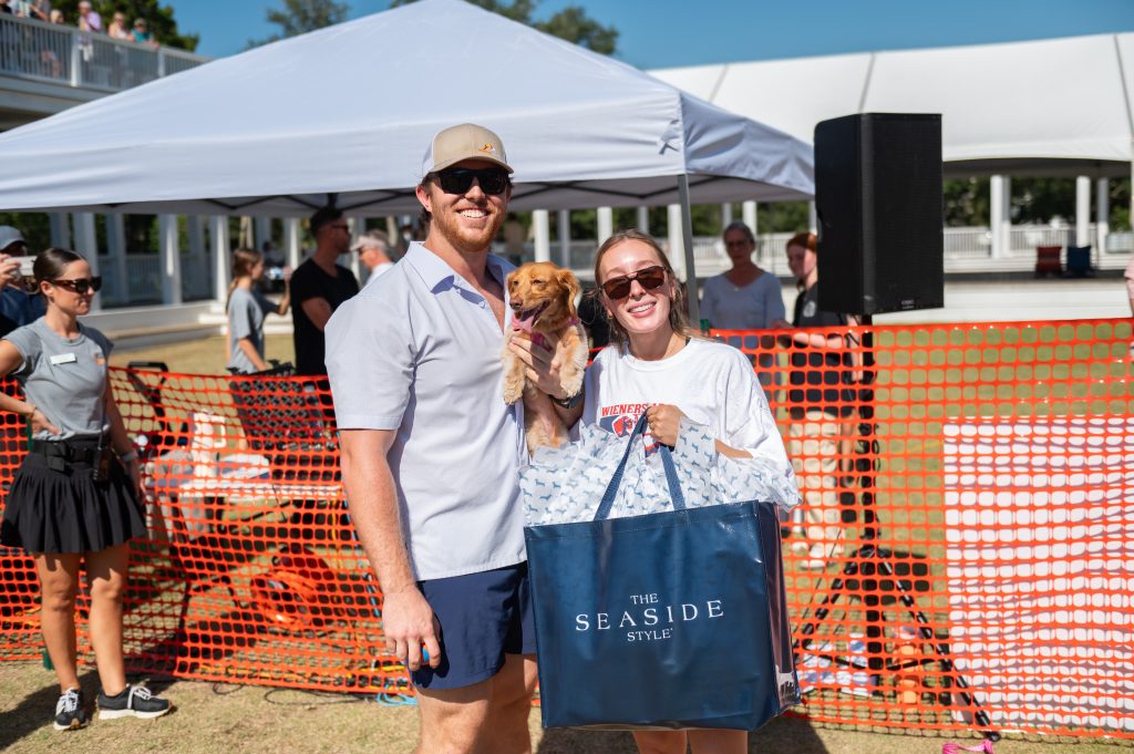 Smiling couple holding a small dog and a Seaside Style bag at an outdoor event.