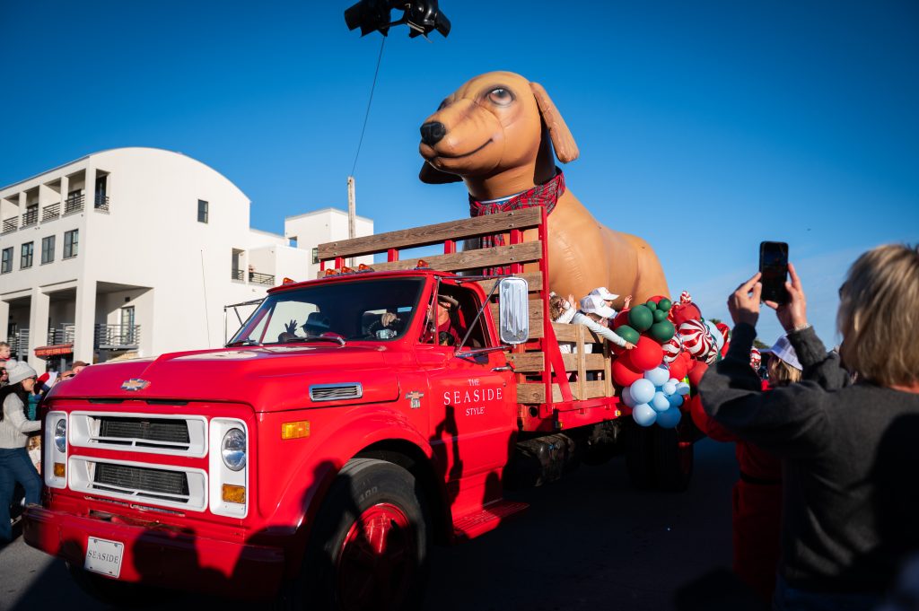 Bright red parade truck carrying large inflatable dog and balloons, surrounded by spectators taking photos.