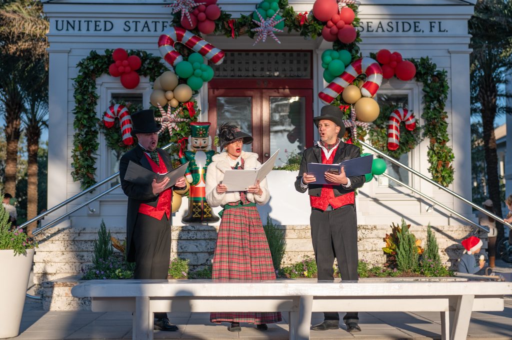 Three carolers in Victorian costumes sing outside a festively decorated building with wreaths and candy cane balloons.