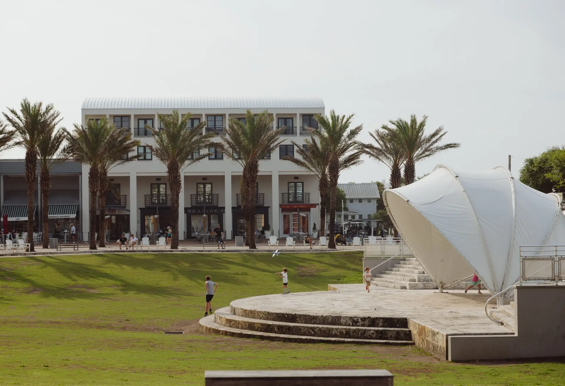 Palm-lined town green with children playing near a white shell-shaped pavilion and shops in the background