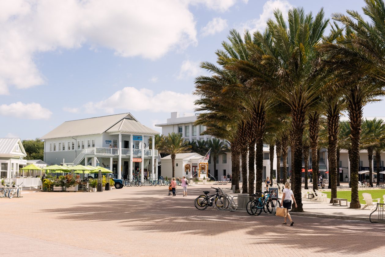 Sunny town square with palm trees, bicycles, and people walking near shops and outdoor seating.