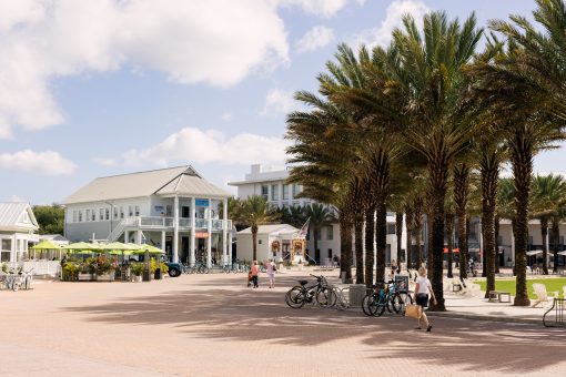 Sunny town square with palm trees, bicycles, and people walking near shops and outdoor seating.