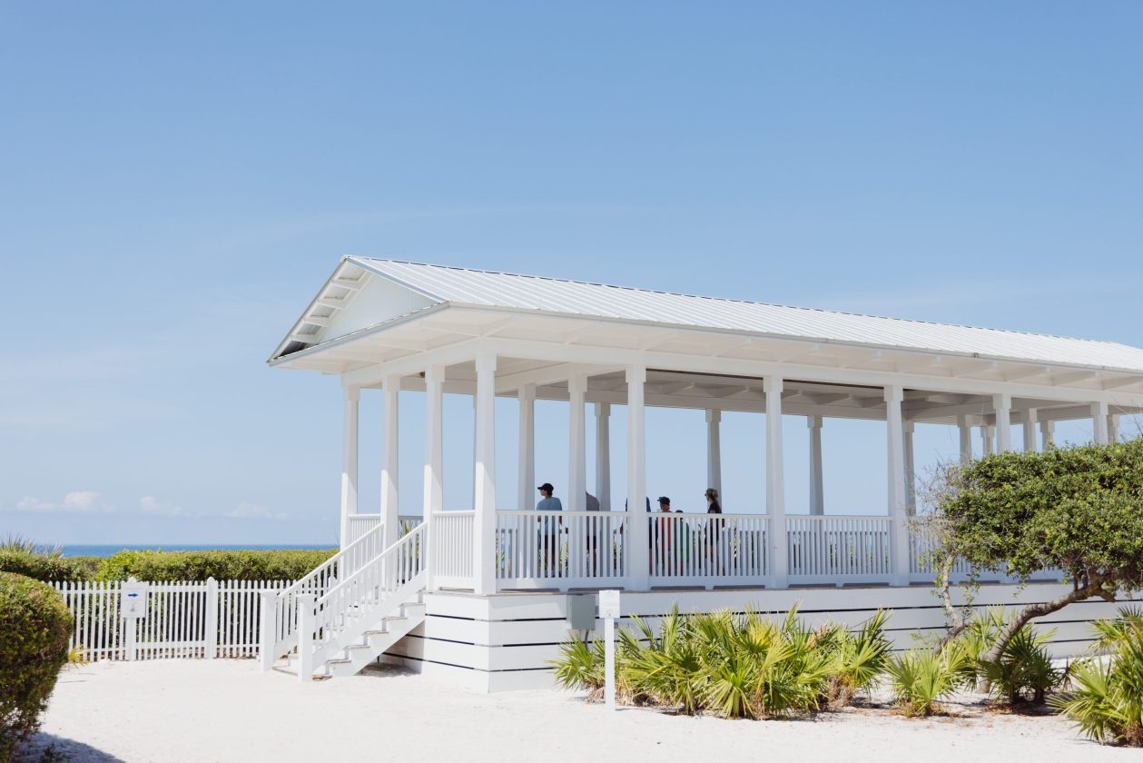 White beach pavilion with people standing inside, overlooking sand and ocean.