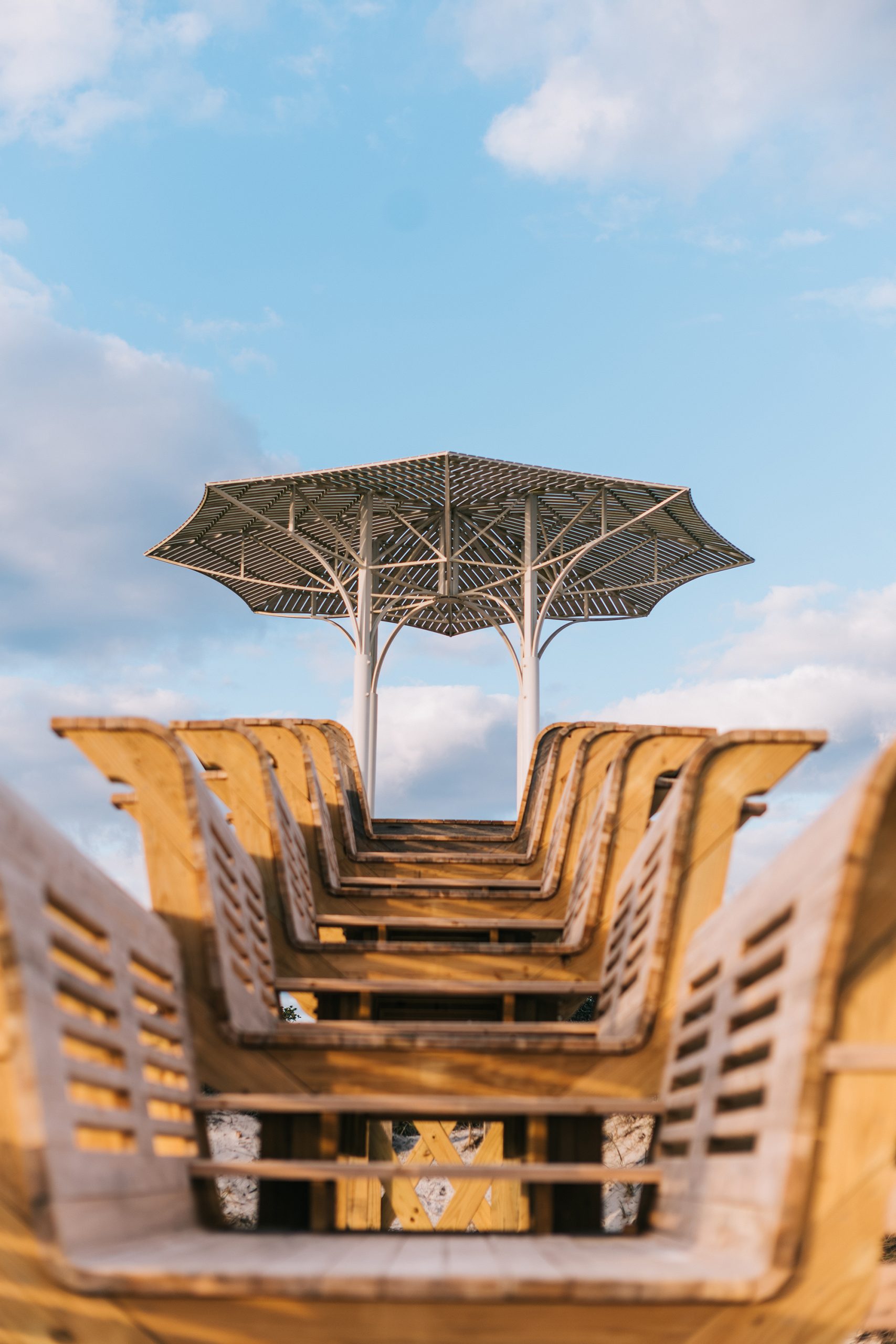 Row of wooden lounge chairs facing a modern geometric shade structure under a blue sky.
