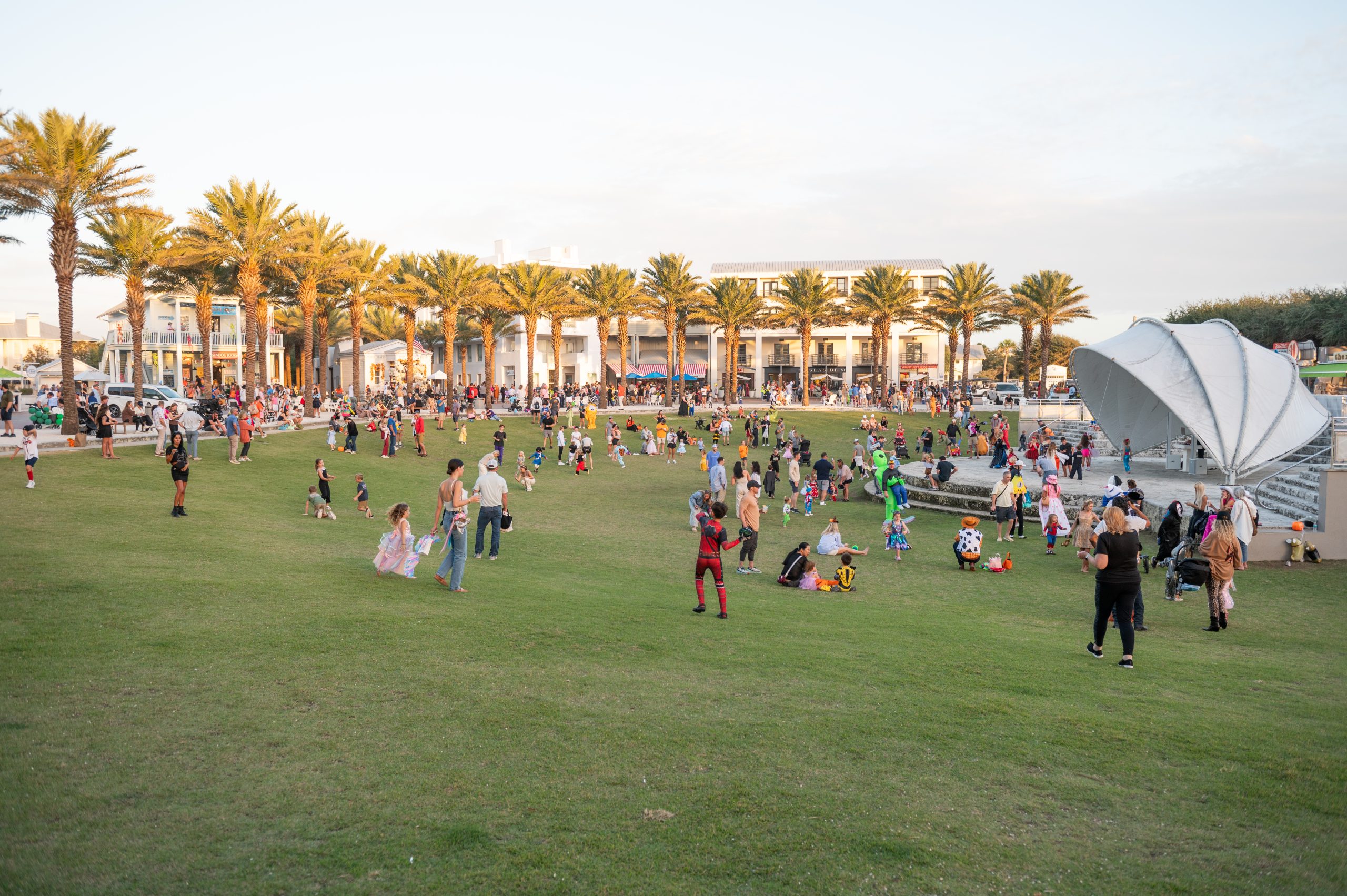 Families gather on a grassy park with palm trees and an outdoor stage at sunset.