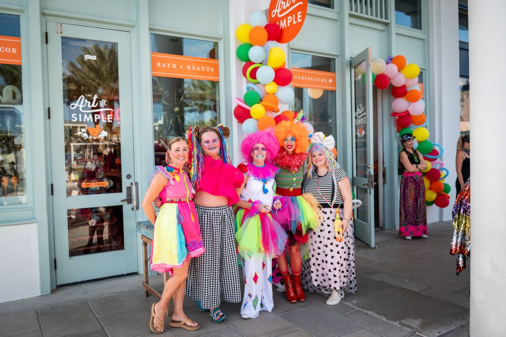 People in colorful, whimsical costumes pose in front of a shop decorated with bright balloon garlands.