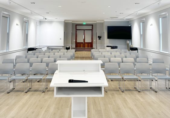 Modern conference room with podium, microphone, and rows of gray chairs facing forward.