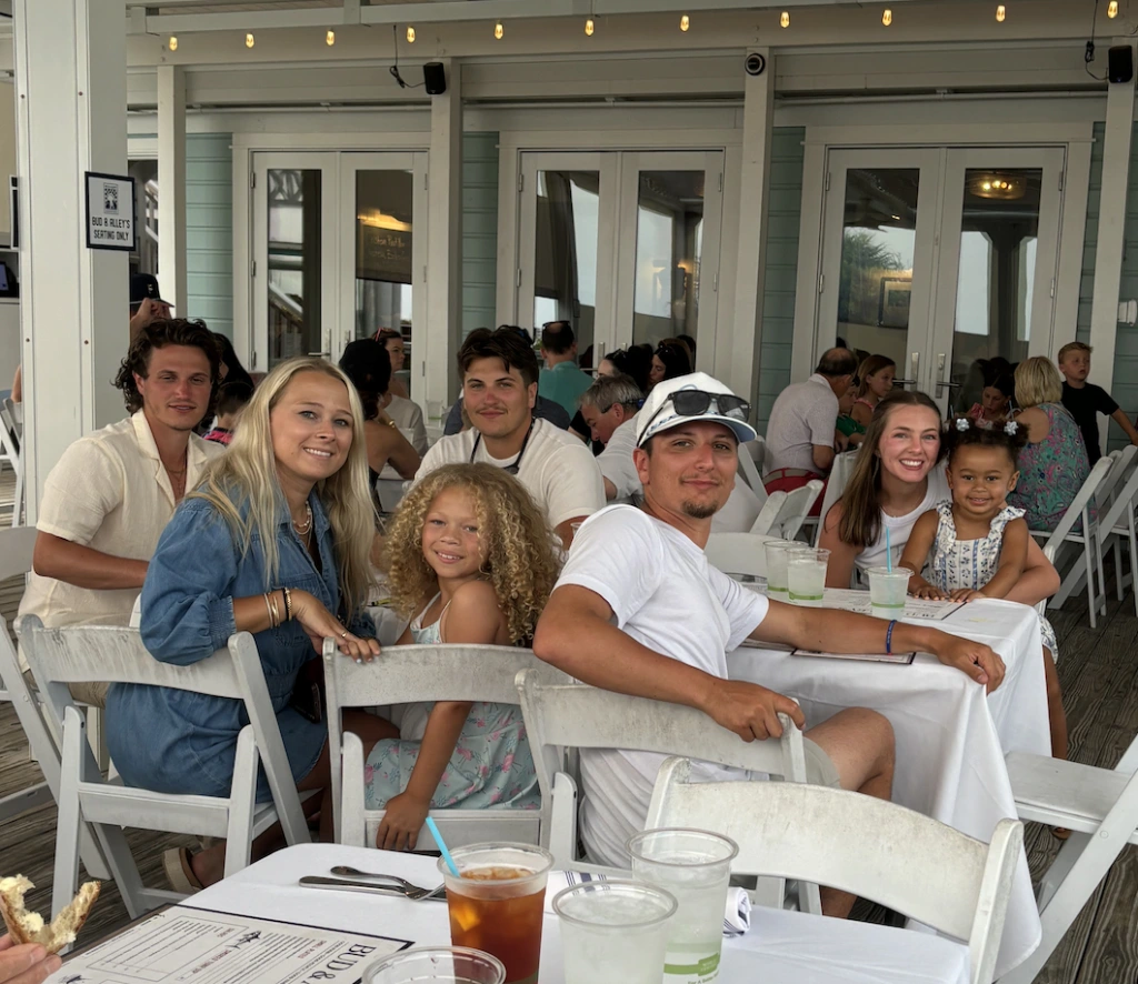 Group of adults and children smiling at a long table during a casual outdoor restaurant gathering with drinks and white chairs.
