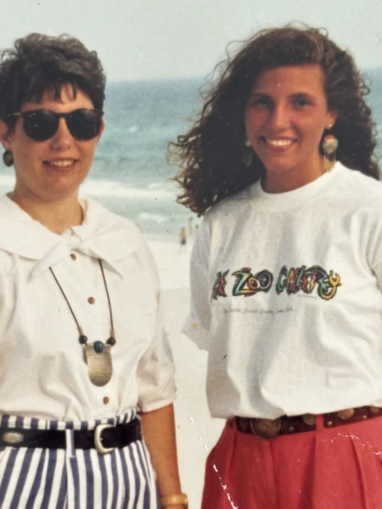 Two women smile at the beach in a vintage photo, wearing casual summer outfits with ocean waves behind them.