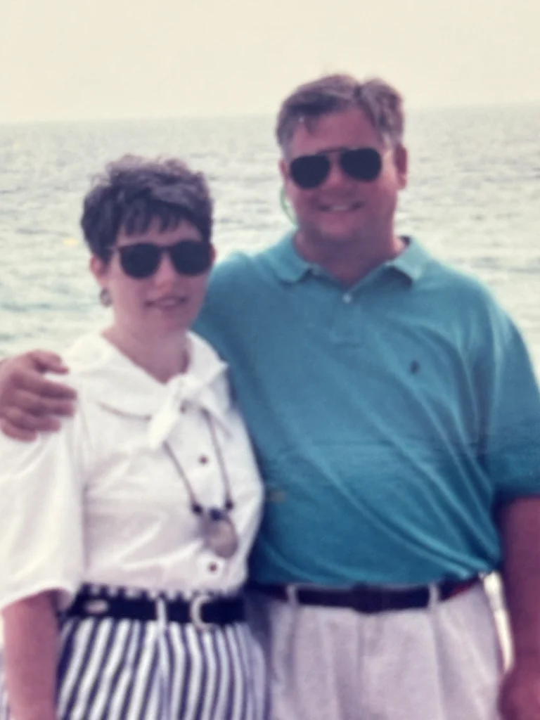 Two adults wearing sunglasses stand close together by the ocean, smiling with waves visible in the background.