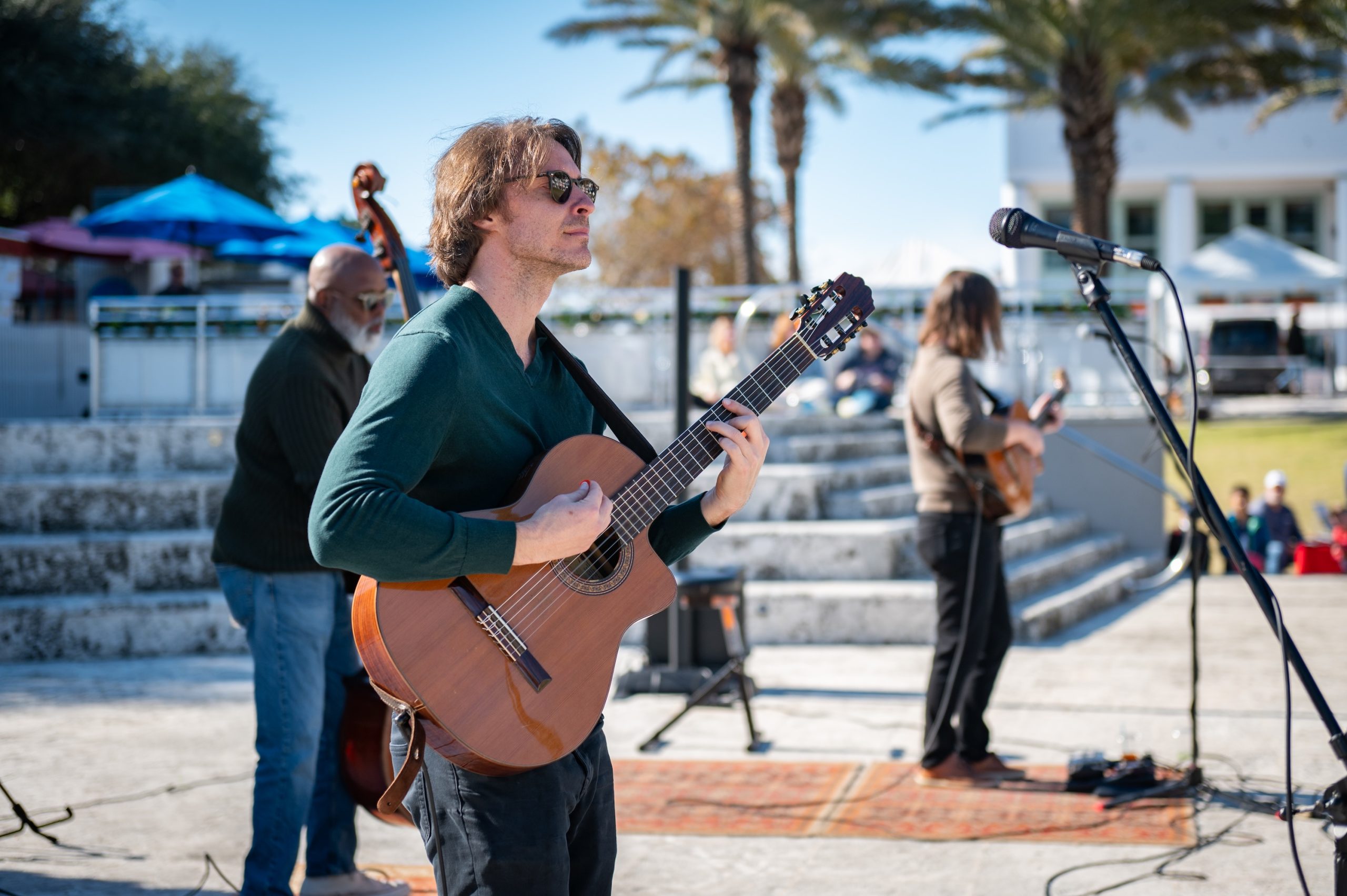 Outdoor musician playing acoustic guitar on sunny stage with bandmates and palm trees.