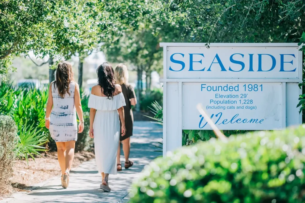 Three women walking along a sunny path beside a Seaside welcome sign surrounded by greenery.