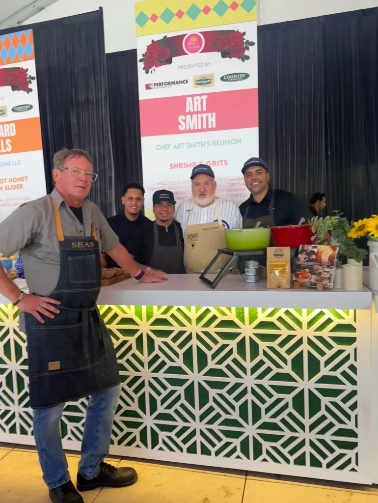 Chef Art Smith and team posing behind a cooking demo counter at a food festival booth.
