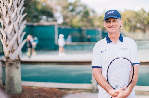 Smiling tennis coach in Seaside uniform holding racket beside outdoor courts and palm tree.