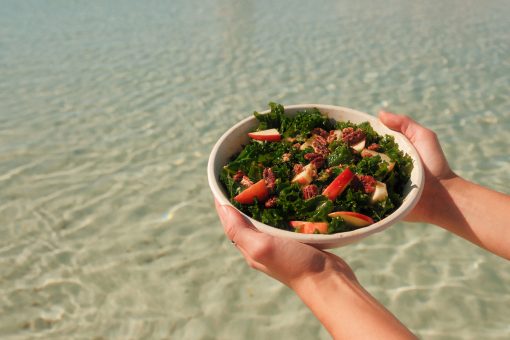 Hands holding kale salad with apples and candied nuts above clear shoreline water.