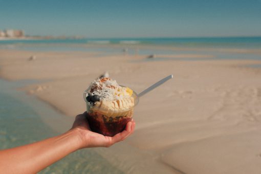Hand holding tropical acai bowl topped with fruit on sandy beach shoreline.