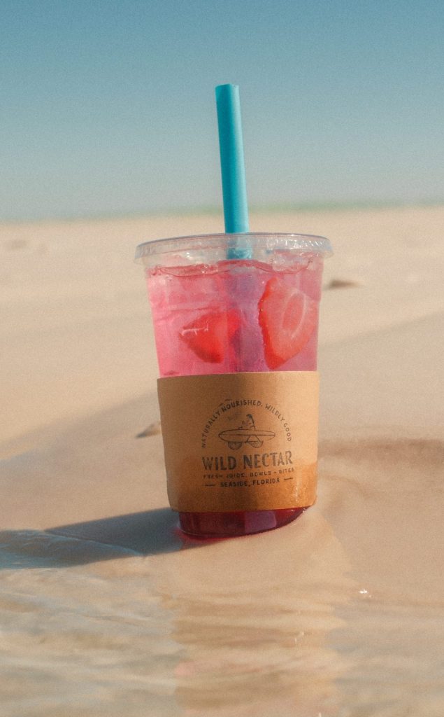 Pink iced drink with fruit slices in plastic cup resting on sunlit beach sand.