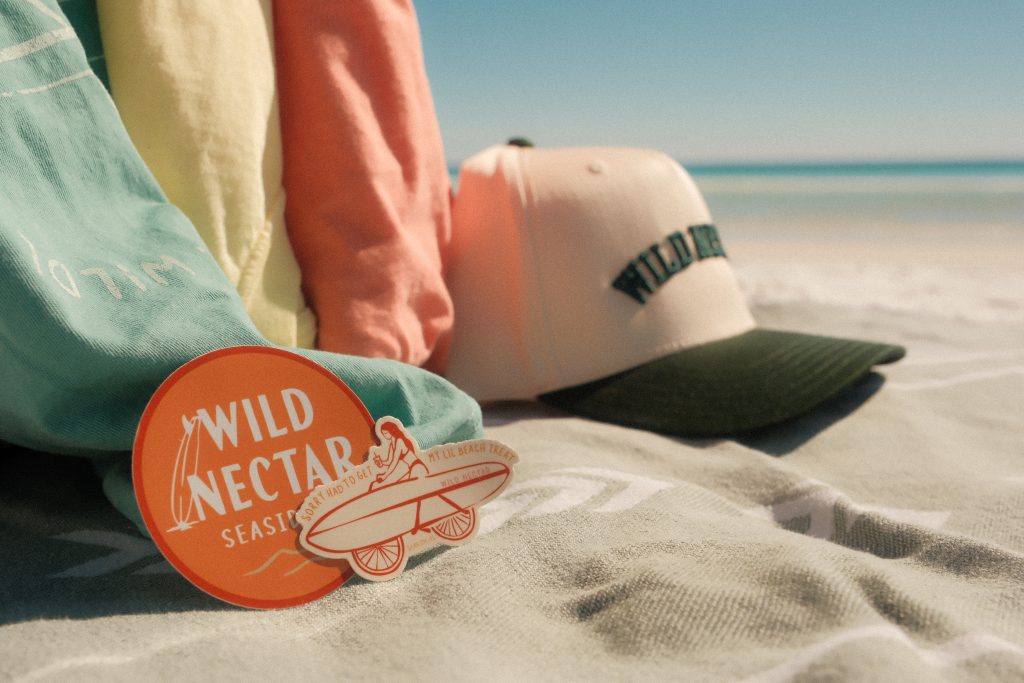Wild Nectar Seaside stickers and pastel towels on a beach blanket, with a branded trucker hat near the ocean.