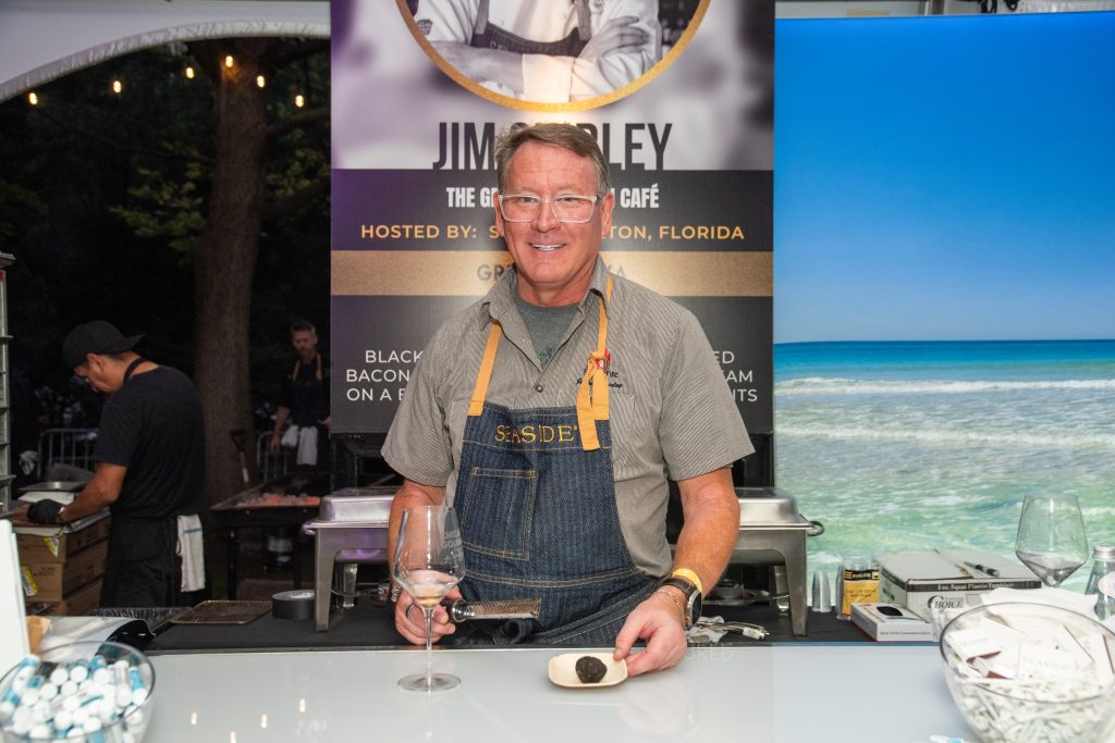 Chef in an apron presents a truffle and wine at an outdoor cooking station during a culinary event.