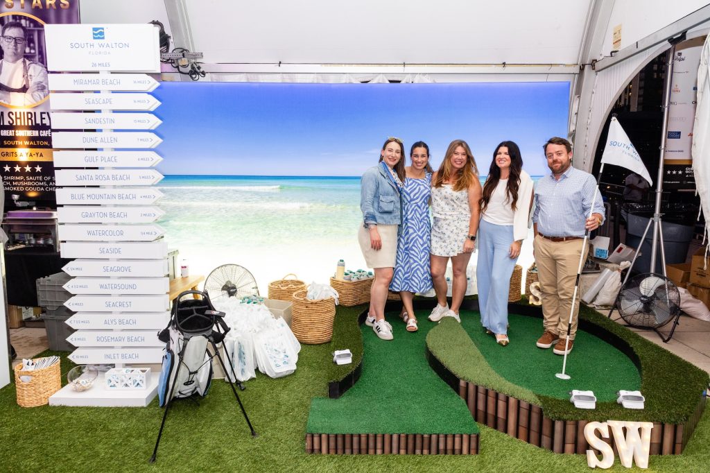 Group posing on mini golf display inside event tent with beach backdrop.