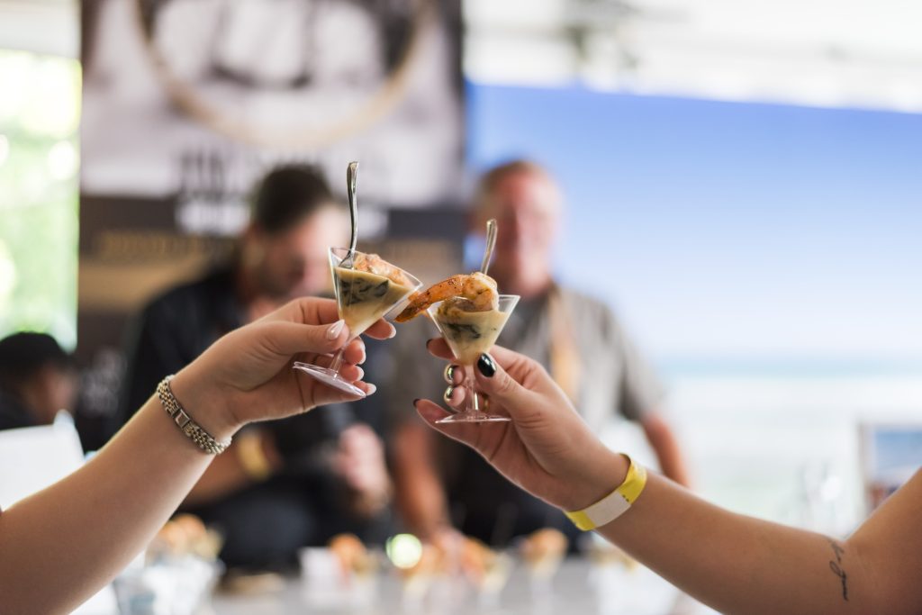 Two hands clink mini dessert parfaits in tiny martini glasses at a lively event.