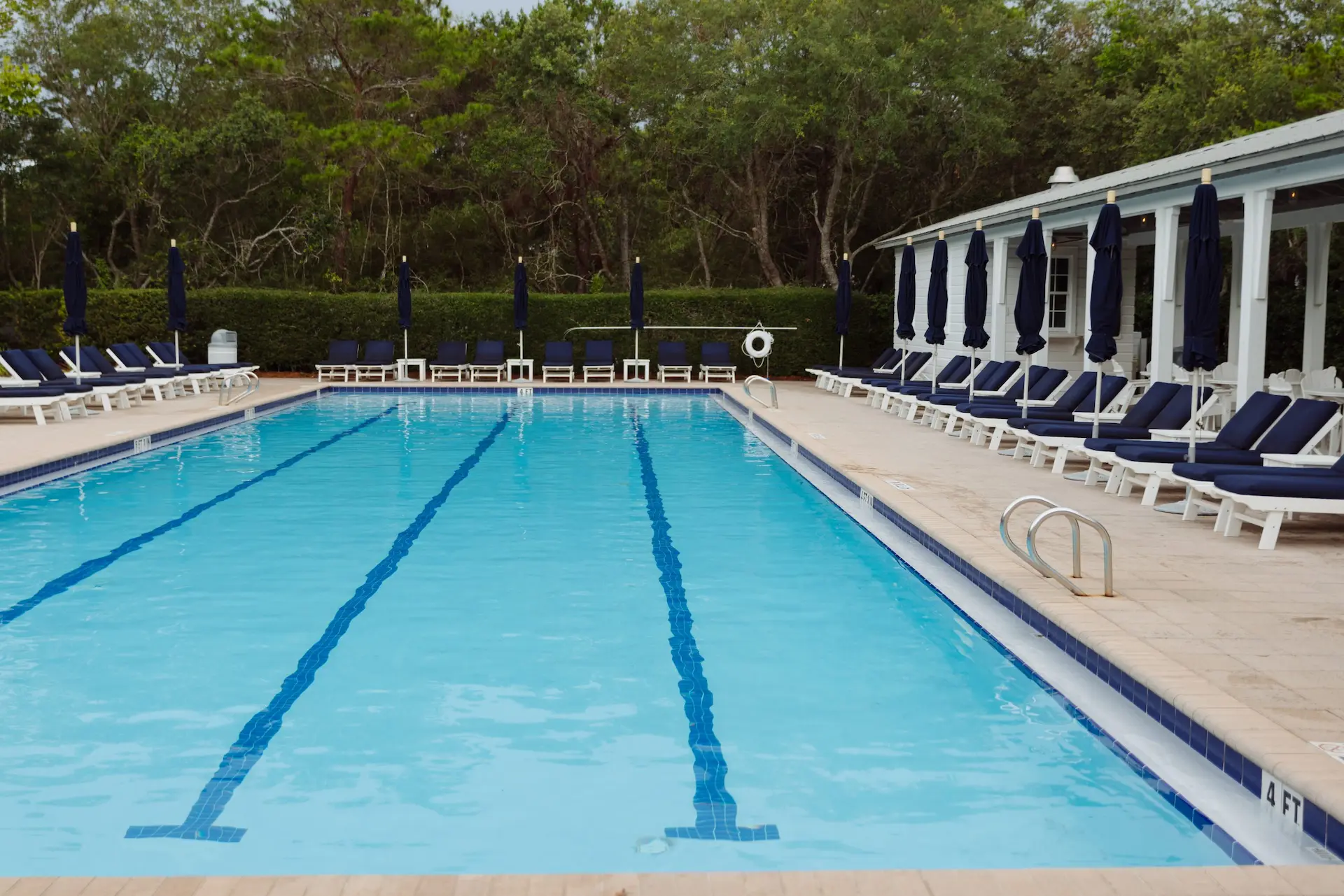 Outdoor swimming pool with blue water, surrounded by lounge chairs and dark umbrellas, trees in background.