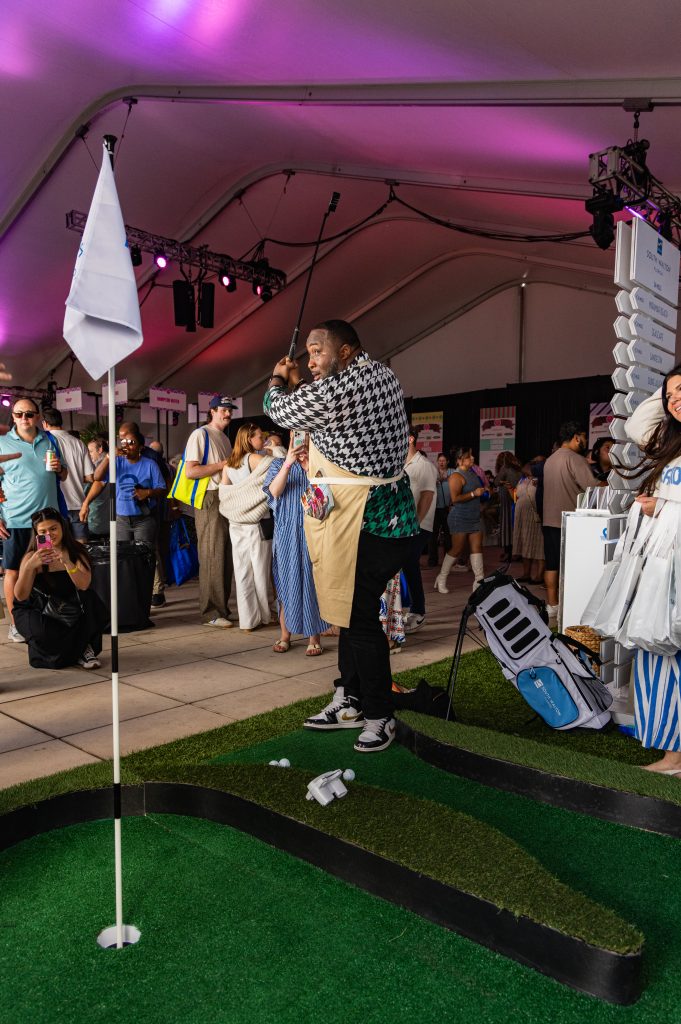 Man takes a golf swing on a mini putting green inside a crowded event tent with onlookers.